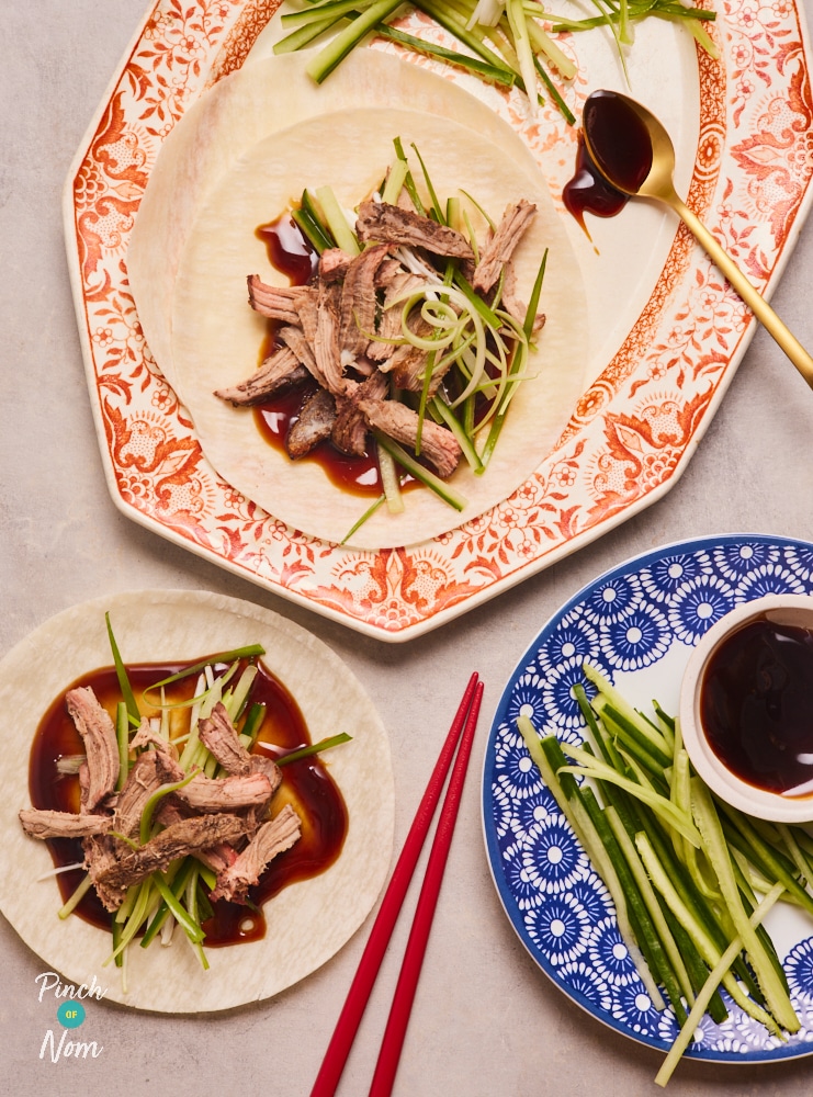 A table is laid with chopsticks, a serving platter of crispy duck, shredded cucumber, spring onions, pancakes and hoisin sauce. Two pancakes are loaded up with fillings, ready to be rolled up and eaten.