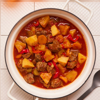 Alt text to add: A large white casserole dish with handles on each side is placed on top of a white, tiled surface. The dish is filled with Pinch of Nom's easy, healthy slow cooker recipe for Pineapple Beef Stew. Juicy chunks of beef, pineapple, carrots, red peppers and celery can be seen nestled in a rich, reddish-brown sauce.