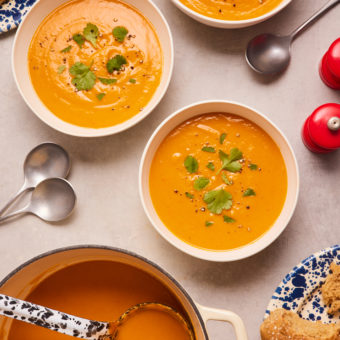 Three bowls of Thai-Spiced Butternut Squash Soup have been filled from a large pan nearby. Each bowl is garnished with fresh coriander, and spoons are place nearby, ready to eat.