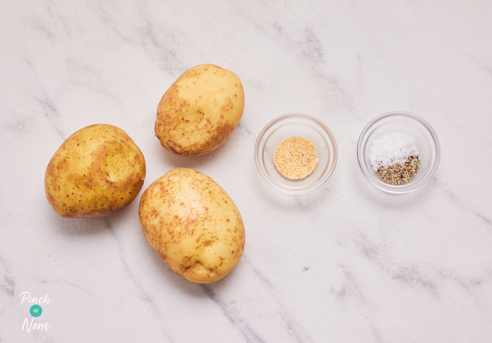 The ingredients for Accordion Potatoes are laid out on a countertop. Three potatoes are nest to two small bowls of salt and pepper and garlic granules.