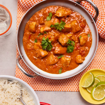 Pinch of Nom's Spicy Coconut and Lime Chicken Curry served in a large pan, covered with a garnish of coriander. Lime wedges are ready to squeeze on a yellow side plate, and basmati rice is ready to be served from a white bowl.