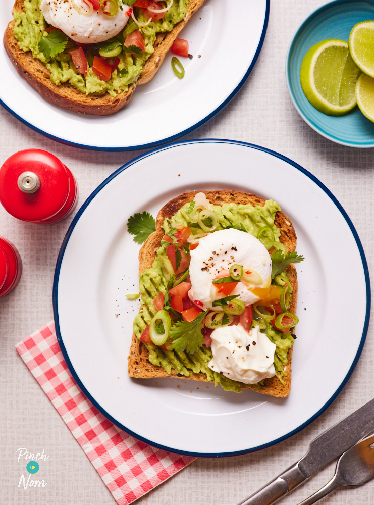 A table is set with two plates of Tex Mex Toast. The toast is topped with smashed avocado, tomato and spring onion salsa, a dollop of fat-free Greek yoghurt and a poached egg. The egg has been cut into and yellow yolk is oozing onto the other toppings.