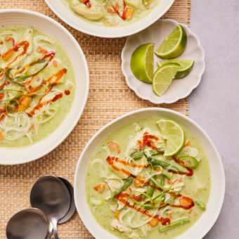 A table is set with three bowls of Pinch of Nom's Thai-Style Green Curry Soup and some sliced limes. The green broth is packed full of chicken, noodles and vegetables, with sriracha sauce drizzled on top. Spoons are laid out nearby, ready to dig in.