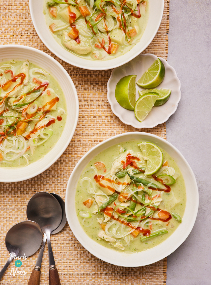 A table is set with three bowls of Pinch of Nom's Thai-Style Green Curry Soup and some sliced limes. The green broth is packed full of chicken, noodles and vegetables, with sriracha sauce drizzled on top. Spoons are laid out nearby, ready to dig in.
