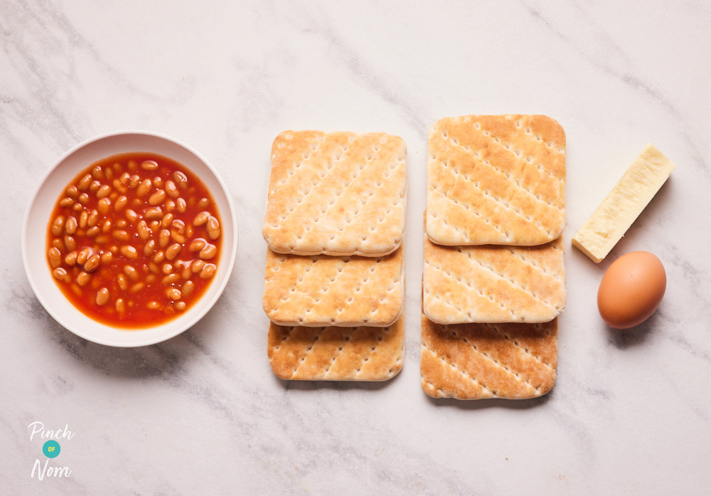 The ingredients for Pinch of Nom's Cheesy Baked Bean Bakes are laid out on a countertop. Alongside baked beans and sandwich thins there is an egg and a small block of reduced-fat Cheddar cheese.