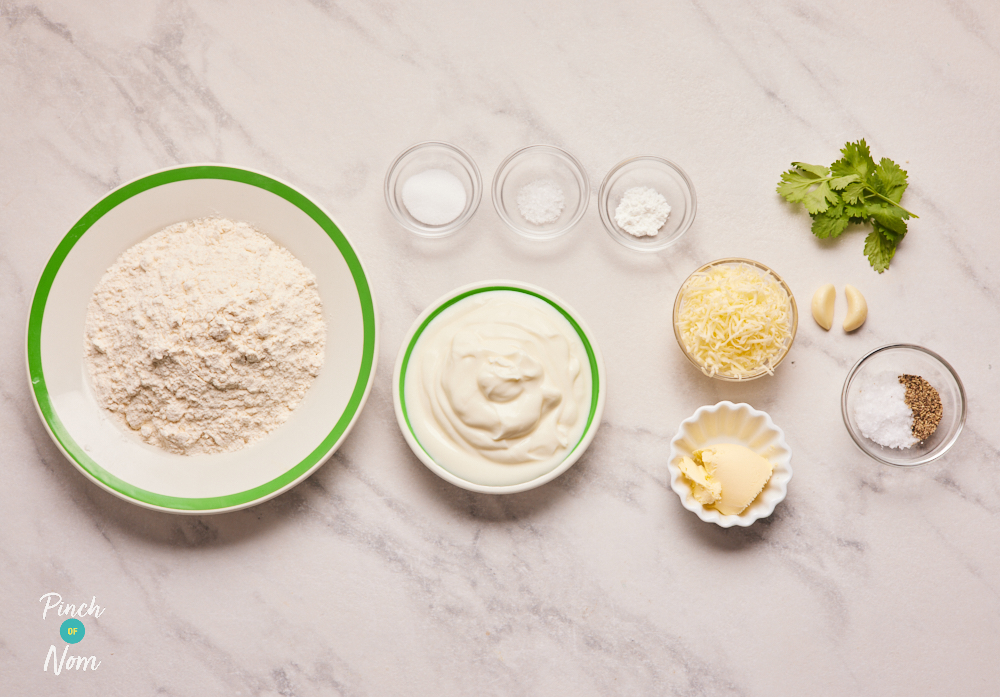 The ingredients for Pinch of Nom's Cheesy Garlic Naan are laid out on a countertop. Alongside flour and fat-free natural yoghurt there is caster sugar, salt, baking powder, reduced-fat spread, garlic cloves, reduced-fat Cheddar cheese and fresh coriander leaves.
