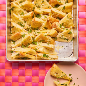 Pinch of Nom's Cheesy Garlic Naan is served on a pink and orange chequered tabletop in a baking tray; each slice is cut into triangles that have been baked until golden brown, topped with cheese and sprinkled with coriander. There are a couple of half-eaten triangles on a pink side plate.