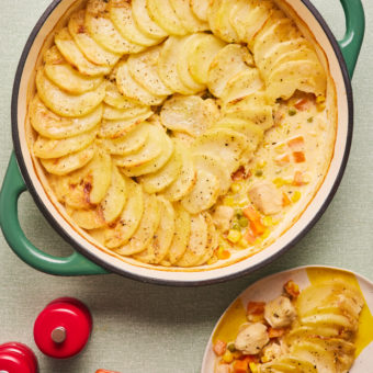 Pinch of Nom's Chicken and Vegetable Hotpot served in a large ovenproof dish with a piece removed where the creamy chicken hotpot is peaking out from underneath the potato topping. A portion lies on a side plate with a spoon waiting to tuck in.