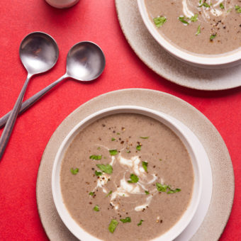 On a red table, two bowls of Pinch of Nom's low-calorie Cream of Mushroom Soup are served. The soup is topped with fresh parsley, and silver soup spoons are nearby, ready to dig in.