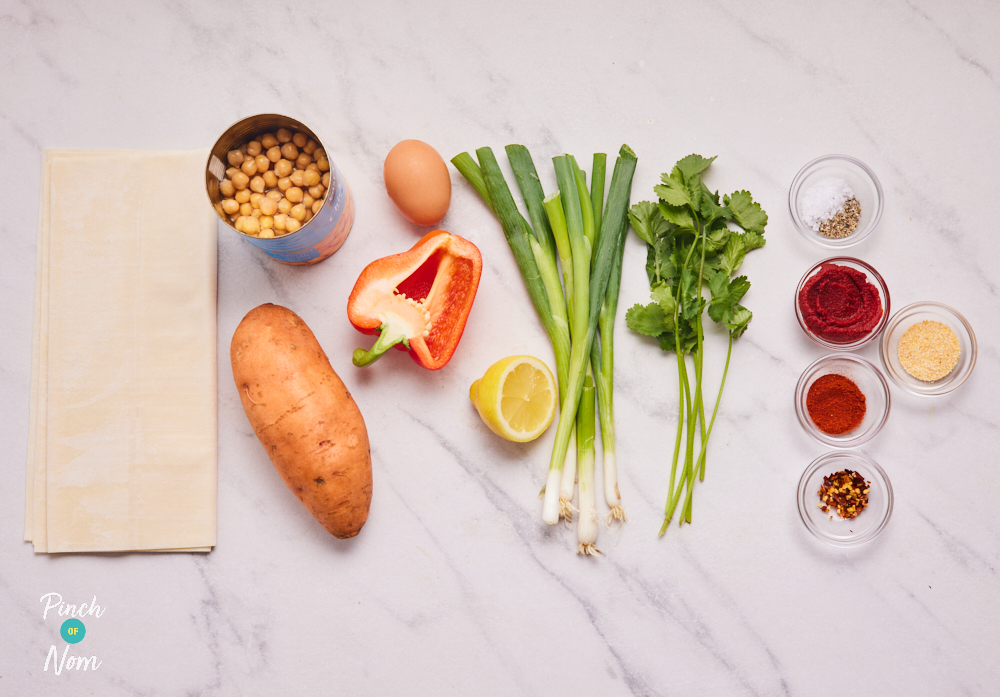 The ingredients for Pinch of Nom's Empanadas are laid out on a countertop. Alongside filo pastry there is a sweet potato, canned chickpeas, an egg, red pepper, lemon, spring onions and a selection of herbs and spices.