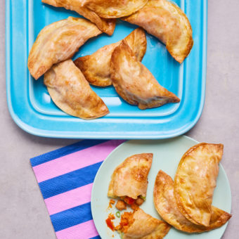 A baking tray of Empanadas is fresh out of the oven, and three Empanadas have been served onto a small pale green plate. The Empanadas are golden brown and one has been cut in half to reveal the vegetable filling.