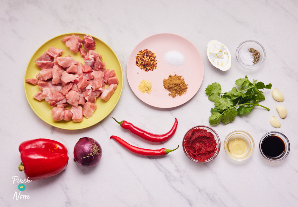 The ingredients for Pinch of Nom's low-calorie Spicy Slow Cooked Pork are laid out on a counter top. There is diced pork, onion, red chillies, herbs, spices, Henderson's relish, stock.