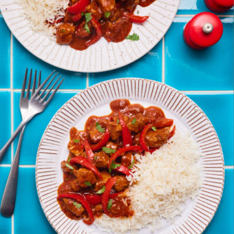 2 portions of Pinch of Nom's Spicy Slow Cooked Pork are plated up on a blue tiled tabletop with forks to the side waiting to dig in. Each portion is served with fluffy basmati rice.