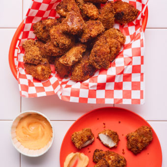 Pinch of Nom's Chipotle Popcorn Chicken is served in a takeaway-style red plastic basket, lined with red and white chequered paper. On a small red plate, a few pieces of popcorn chicken have been served with some sauce. An additional pot of sauce is nearby, ready for the chicken to be dipped in.
