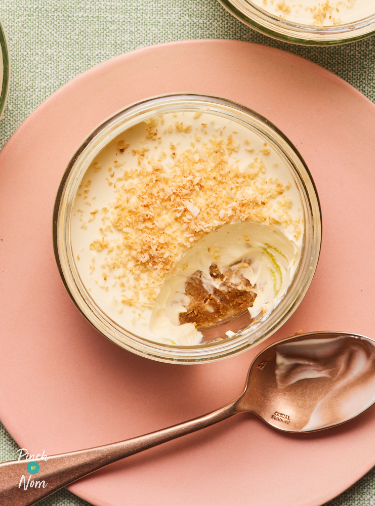 A close-up of one of Pinch of Nom's Creamy Lime and Coconut Pots served in a glass ramekin, with a spoon to the side that's waiting to tuck in. A spoonful has already been scooped out, revealing the Biscoff layers beneath the creamy top.