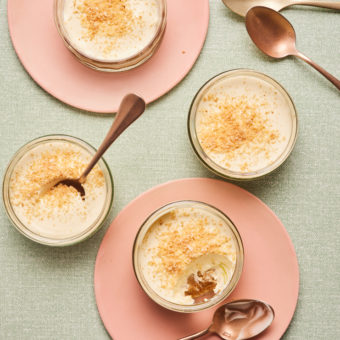 Four of Pinch of Nom's Creamy Lime and Coconut Pots are served on pink plates inside individual glass ramekins, with spoons waiting to tuck in. A spoonful has already been scooped out of one of the puddings, revealing the Biscoff layers beneath the creamy top.