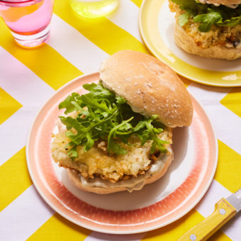 Two of Pinch of Nom's Crispy Fish Burgers are piled high on colourful plates with tartare sauce, crispy-coated cod fillets and rocket.