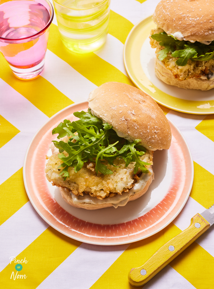 Two of Pinch of Nom's Crispy Fish Burgers are piled high on colourful plates with tartare sauce, crispy-coated cod fillets and rocket.