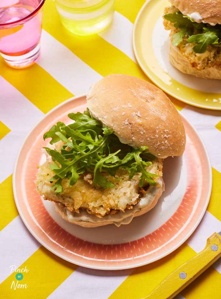 A close-up image of two of Pinch of Nom's Crispy Fish Burgers are piled high on colourful plates with tartare sauce, crispy-coated cod fillets and rocket.