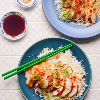 Two blue plates are served with Pinch of Nom's Miso Chilli Chicken. The sliced chicken breasts are arranged on a bed of jasmine rice, garnished with sesame seeds, finely sliced chillies and spring onions. Green chopsticks are balanced on the plate, ready to dig in.