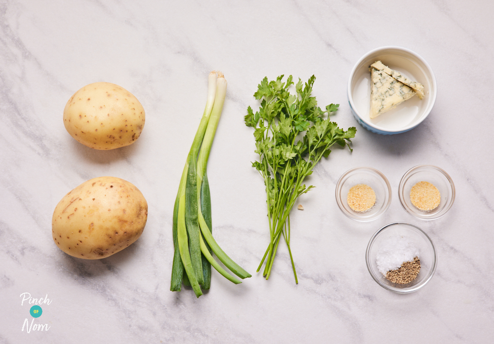 The ingredients for Pinch of Nom's Stilton Roast Potatoes recipe are laid out on a countertop. There are two potatoes, spring onions, fresh parsley, Stilton cheese, garlic granules, onion powder and salt and pepper.