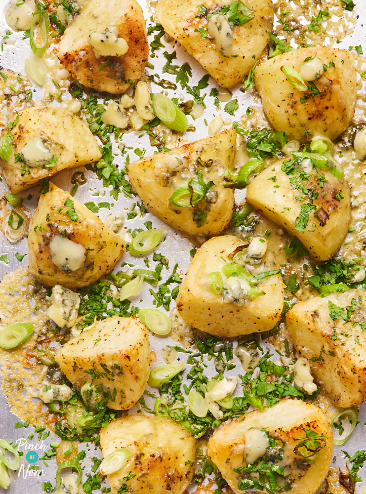 A close up photo of Pinch of Nom's Stilton Roast Potatoes. The potatoes are golden and crispy at the edges, covered with melted Stilton cheese, sliced spring onions and finely chopped parsley.