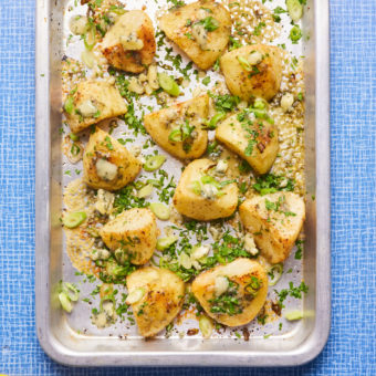 Pinch of Nom's Stilton Roast Potatoes are fresh from the oven, on a silver baking tray. The potatoes are golden and crispy at the edges, covered with melted Stilton cheese, sliced spring onions and finely chopped parsley.