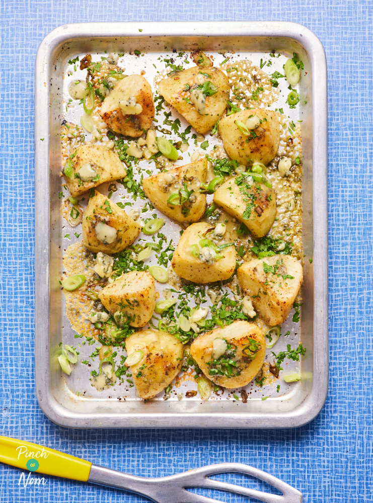 Pinch of Nom's Stilton Roast Potatoes are fresh from the oven, on a silver baking tray. The potatoes are golden and crispy at the edges, covered with melted Stilton cheese, sliced spring onions and finely chopped parsley.