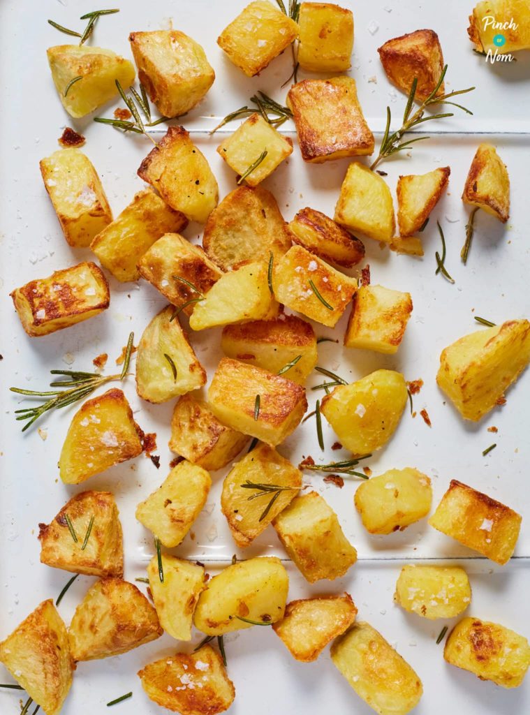 Pinch of Nom's crispy Air Fryer Roast Potatoes are scattered on a white countertop. They are crunchy and golden, with flaky sea salt and sprigs of fresh rosemary scattered over the top.