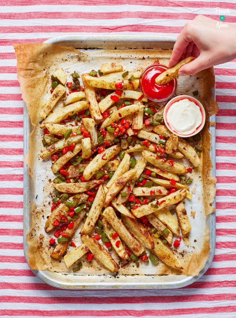A baking tray lined with greaseproof paper is filled with Pinch of Nom's Air Fryer Salt and Pepper Chips. The golden chips are mixed with finely diced red and green peppers and salt and pepper seasoning. A small pot of ketchup and a small pot of mayonnaise are served alongside the chips, and a hand holds one chip, dipping it into the ketchup.
