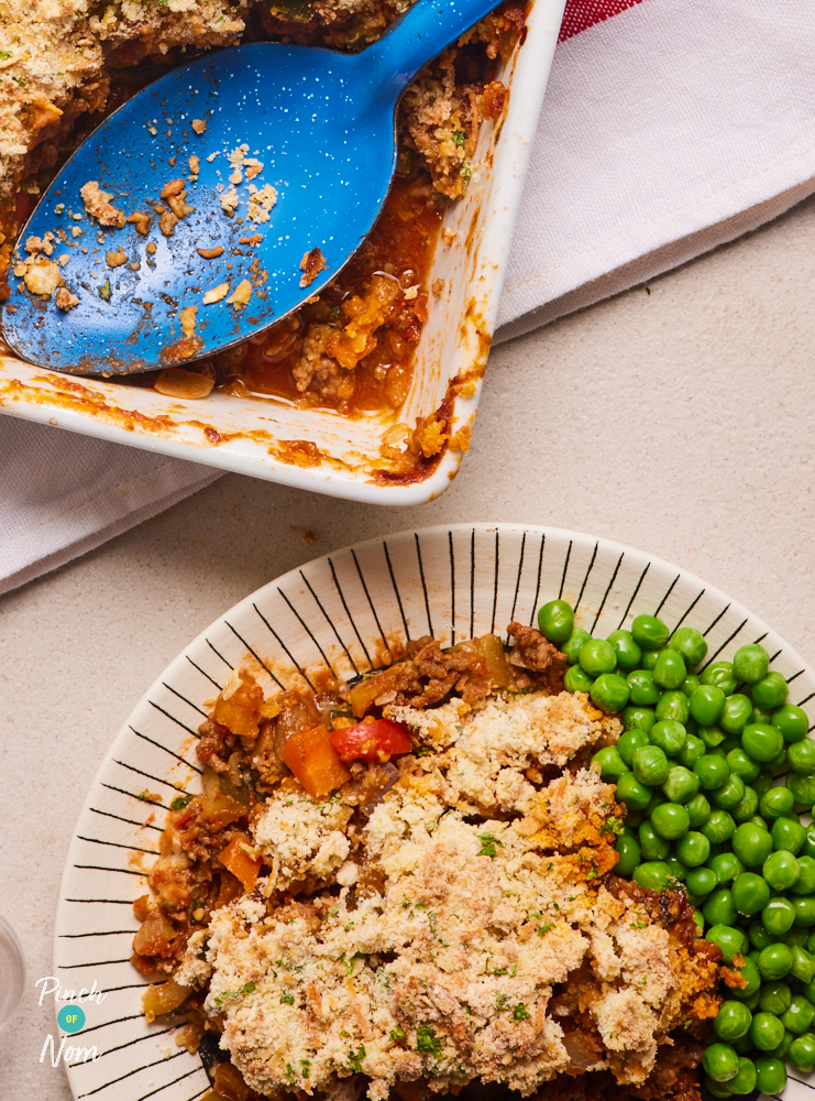 A close up of a plate of Pinch of Nom's Beef Crumble has been spooned onto a plate from an oven dish nearby. A blue serving spoon is resting in the oven dish. The Beef Crumble is golden brown on top, served with vibrant green garden peas.