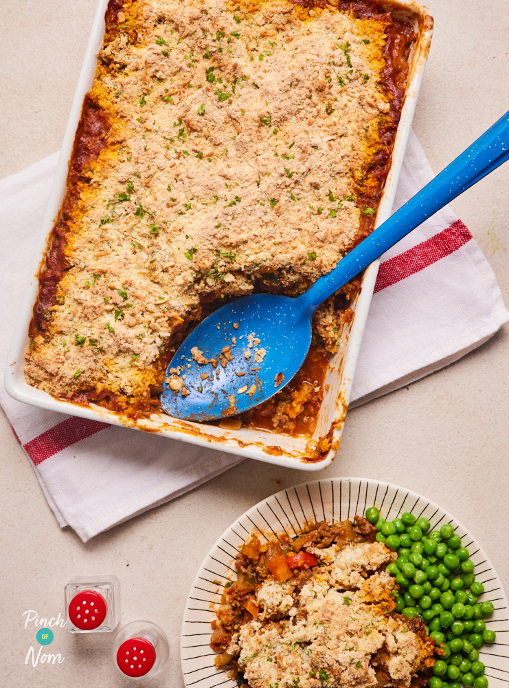 A plate of Pinch of Nom's Beef Crumble has been spooned onto a plate from an oven dish nearby. A blue serving spoon is resting in the oven dish. The Beef Crumble is golden brown on top, served with vibrant green garden peas.