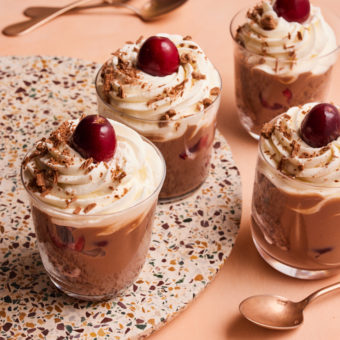 Pinch of Nom's Black Forest Trifles are served in individual glass dishes. Inside the dish you can see the layers of chocolate Swiss roll, and chocolate custard, topped with a swirl of aerosol cream and a fresh juicy cherry.