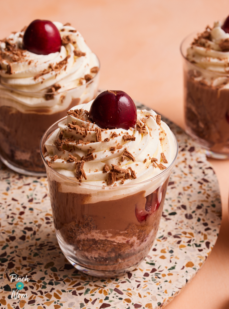 A close-up shot of one of Pinch of Nom's Black Forest Trifles, served in individual glass dishes. Inside the dish you can see the layers of chocolate Swiss roll and chocolate custard, topped with a swirl of aerosol cream and a fresh juicy cherry.