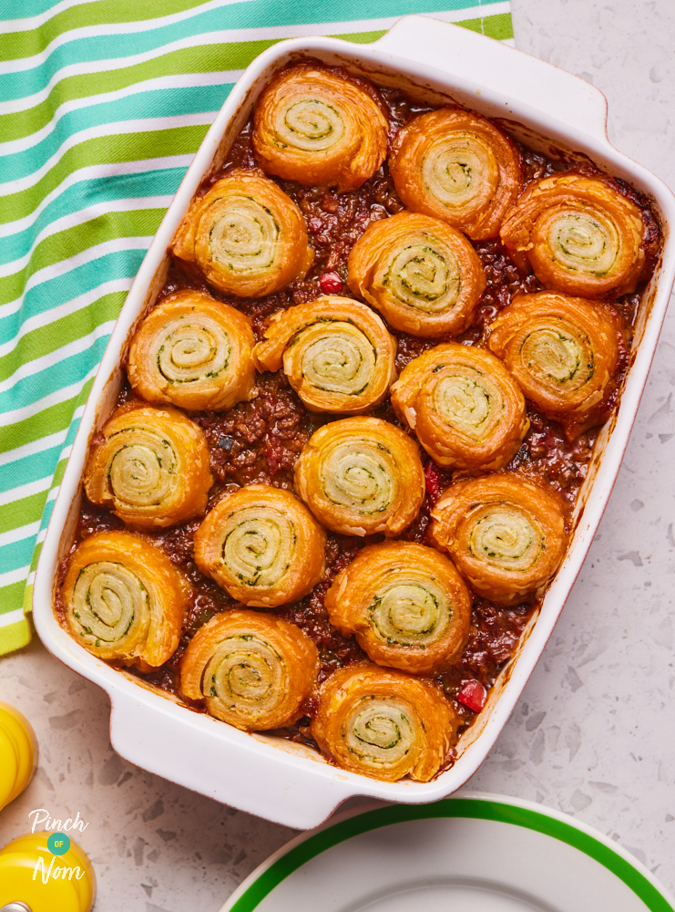 Pinch of Nom's Bolognese and Garlic Swirl Pie is served, fresh from the oven, in a large ovenproof dish on a table set with salt and pepper shakers. The garlic swirls are an eye-catching golden brown on top, evenly covering the bolognese filling.