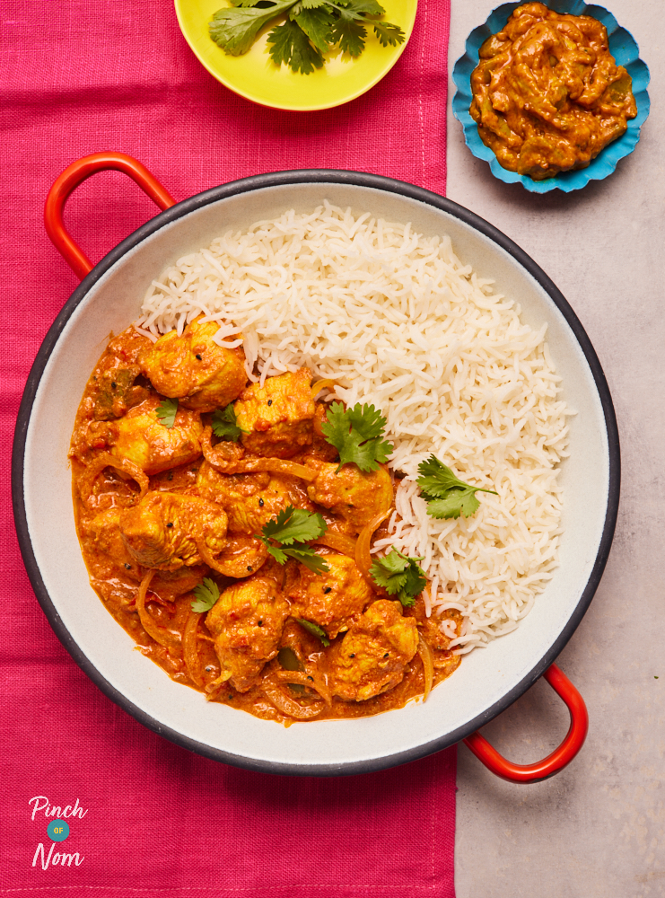 Pinch of Nom's Chicken and Mango Pickle Curry is served in a large round dish with red handles. The curry takes up one half of the dish, and fluffy white basmati rice is on the other side.