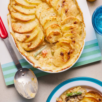 Pinch of Nom's Creamy Vegetable Hotpot is served fresh from the oven, in an oval-shaped oven dish. The potato topping is golden and crisp, and a spoonful has been dished up onto a dinner plate. The vegetable filling is creamy and colourful.