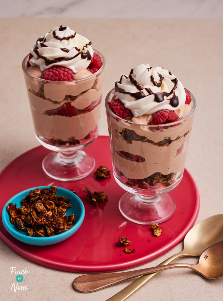 Two glasses of Pinch of Nom's Raspberry Mocha Granola pots are served, topped with a swirl of cream, raspberries and a drizzle Sweet Freedom Choc Shot. Extra granola waits to the side to be sprinkled on top.
