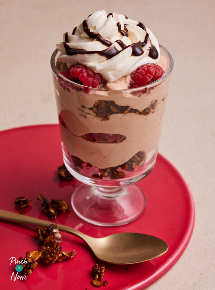 A glass of Pinch of Nom's Raspberry Mocha Granola Pots is served on a red plate, topped with a swirl of cream, raspberries and a drizzle Sweet Freedom Choc Shot. Extra granola waits to the side to be sprinkled on top.