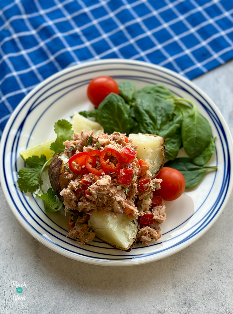 A plate of Pinch of Nom's Air Fryer Fluffy Jacket Potatoes with Spicy Tuna is served on a white tabletop, topped with a garnish of chopped chillies and a colourful side salad.