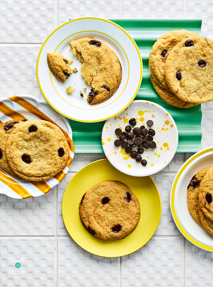 A table is set with small plates of Pinch of Nom's Air Fryer Freezer Cookies from the Pinch of Nom: Air Fryer cookbook, with an extra side plate of chocolate chips. One of the cookies is already half-eaten!