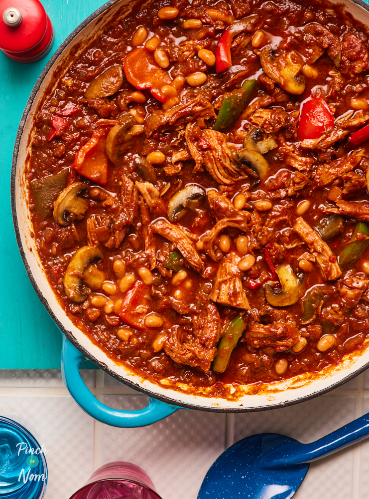 On a kitchen counter, a large casserole dish is resting on a bright blue chopping board. The dish contains Pinch of Nom's BBQ Gammon Stew, fresh from the oven. The stew is packed with colourful vegetables, baked beans and shredded gammon, in a deep red tomatoey sauce. A serving spoon is nearby, ready to dish up dinner.