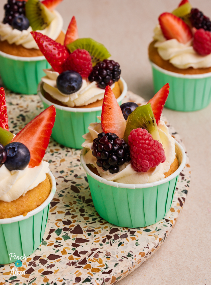 A close-up of Pinch of Nom's Fruity Cupcakes served in colourful cases on a speckled serving plate topped with strawberry, kiwi, blueberry and raspberry slices.