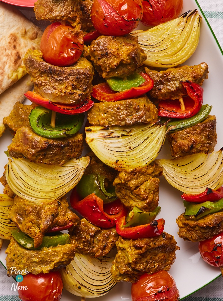 A close up of Pinch of Nom's Indian-Spiced Lamb Kebabs. The spiced meat is threaded onto a wooden skewer, interspersed with chunks of red and green peppers, onion and cherry tomatoes.