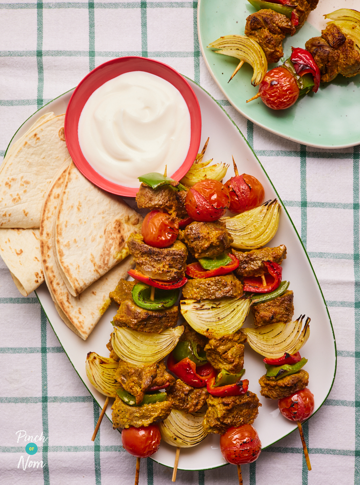 A table is set with a white check-print tablecloth. A platter of Pinch of Nom's Indian-Spiced Lamb Kebabs is in the centre of the table. The lamb skewers are colourful, with pops of cherry tomatoes and green and red peppers. Tortilla wraps have been folded into triangles and arranged on the platter, next to a small red bowl filled with fresh yoghurt. To the side, a small green plate holds 2 of the kebabs.