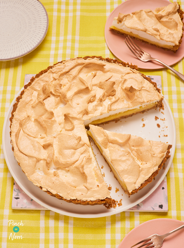 A table is set with a yellow chequered-pattern tablecloth. On a large white plate, Pinch of Nom's Lemon Meringue Pie is being sliced into servings. One slice has already been placed onto a small pink plate and another triangular slice is ready to be served.