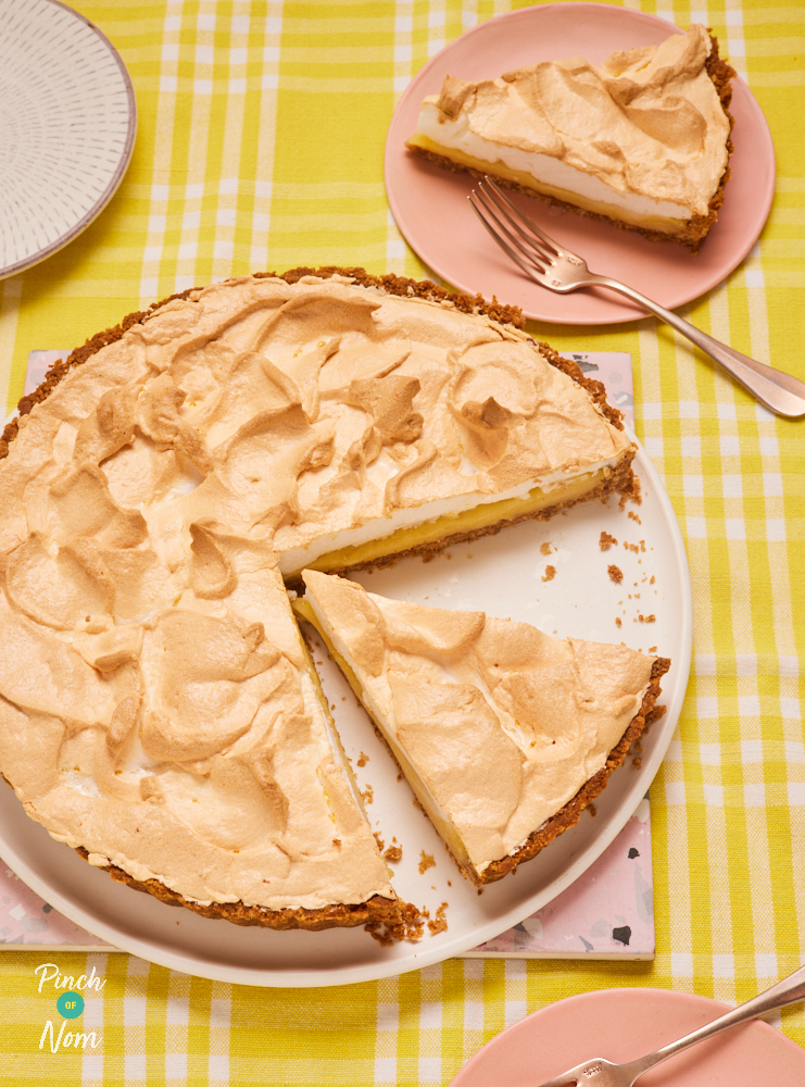 A table is set with a yellow chequered-pattern tablecloth. On a large white plate, Pinch of Nom's Lemon Meringue Pie is being sliced into servings. One slice has already been placed onto a small pink plate and another triangular slice is ready to be served.