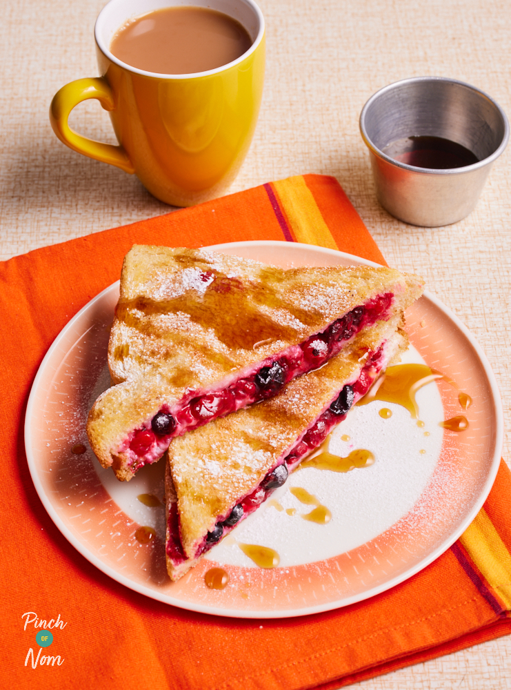 Pinch of Nom's Berry Toastie is served on a white and orange plate, on top of a bright orange place mat. A cup of tea waits nearby, and a small metal pot of maple syrup. The toastie has been cut into two triangles, and one rests on top of the other. The mixed berry filling can be seen in-between slices of golden toast, and icing sugar has been sprinkled over the top. The toastie is drizzled with maple syrup.