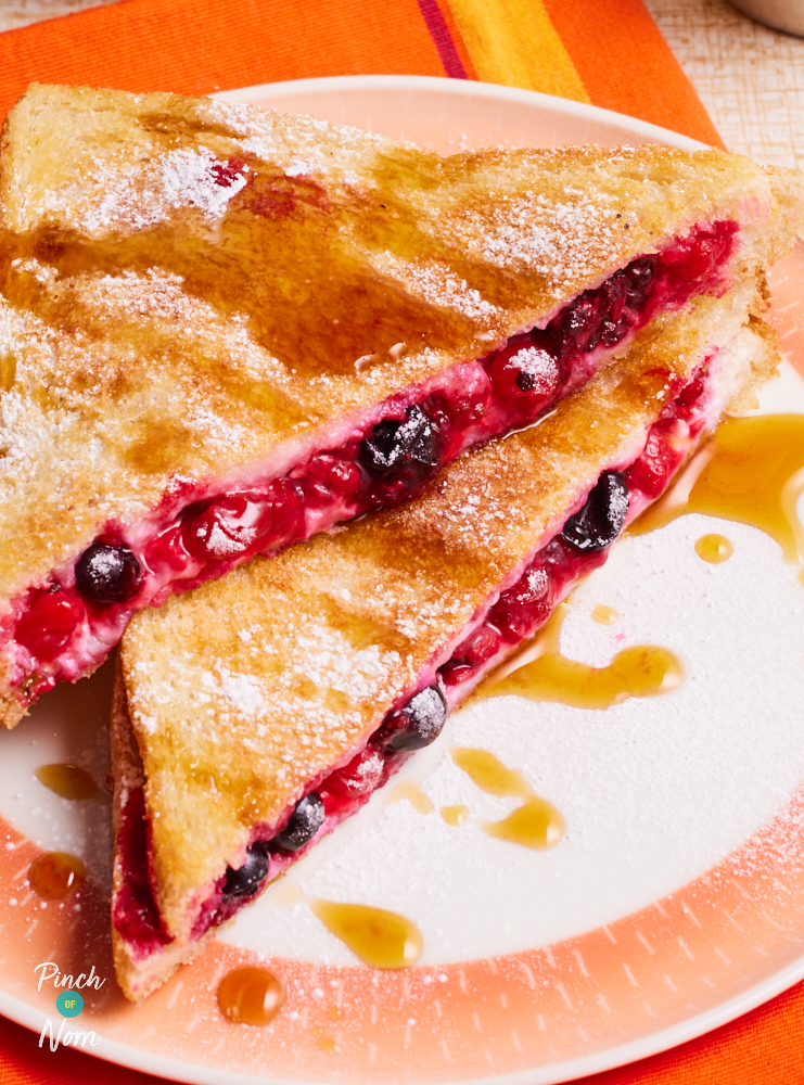 A close-up shows Pinch of Nom's Berry Toastie served on a white and orange plate. The toastie has been cut into two triangles, and one rests on top of the other. The mixed berry filling can be seen in-between slices of golden toast, and icing sugar has been sprinkled over the top. The toastie is drizzled with maple syrup.