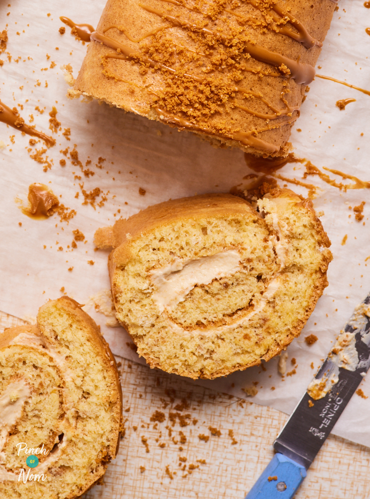 A close-up of Pinch of Nom's Biscoff Swiss Roll served drizzled with melted Biscoff spread and a sprinkling of crushed Biscoff biscuits on top. Two slices have been cut from the end, revealing a swirl of the homemade Biscoff filling inside.